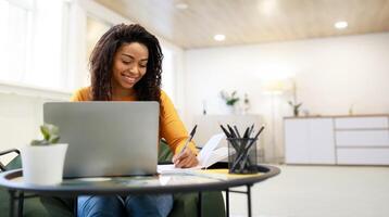 Smiling young black woman sitting at desk working on laptop taking notes in notebook, happy millennial female study watching webinar using computer writing check list, banner panorama, free copy space photo