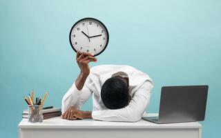 Tired african american businessman sitting at desk with pc laptop, holding clock and putting head on hand, feeling exhausted over blue studio background photo