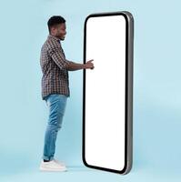 Full Body Length Back View Of Black Man Using Big Smartphone With Blank White Screen Touching Huge Display Panel With Finger, Cheerful Guy Standing On Blue Background, Ordering Food Delivery, Mock Up photo