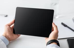 Online digital touch screen with ad, offer and website or medical blog. Young businessman or doctor in coat with smart watch uses tablet with blank screen on white table with notepads, close up photo