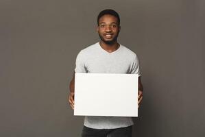 Picture of young smiling african-american man holding white blank board on grey background, copy space photo