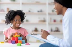 Friendly attractive african american woman child development specialist having session with cheerful little girl, sitting at table, playing with colorful wooden bricks, having conversation and smiling photo