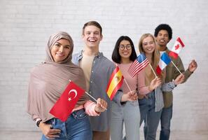 Modern exchange program and language learning. Happy young international students looking at camera and holding flags of their countries, on white brick wall background, studio shot, copy space photo