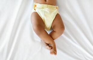 Bottom Part Of Black Infant Baby Wearing Reusable Diaper Lying On Bed, Top View Of Unrecognizable Little African American Boy Or Girl Relaxing On White Linens In Bedroom, Cropped Shot With Copy Space photo