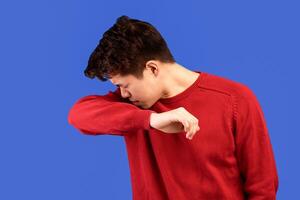 Stop Virus And Germs Spreading Concept. Portrait of young asian man wearing red sweater coughing in his elbow, sneezing in correct way, isolated over ble studio backgound, banner. Health Care photo