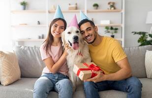 Happy diverse couple celebrating their pet dog's birthday, sitting on sofa with gift box, wearing festive hats at home. Adorable golden retriever having b-day with owners, enjoying party photo