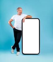 Cheerful smiling mature man looking at giant big cell phone, leaning on empty white screen template panel, standing on blue studio wall. Mock up for app or website, free copy space, full body length photo
