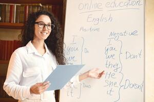 Lecture And Online Education Concept. Portrait of smiling positive young lecturer wearing eyeglasses teaching English language, pointing at grammar rules on board, holding clipboard, explaining photo