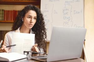 Distant Remote Work. Portrait of serious woman in earbuds having conference on computer, showing paper document to webcam with pen, giving presentation or online class to students photo