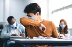 Stop Spreading Virus Concept. Asian male student couching in his elbow, sneezing in sleeve during lecture in university. Guy wearing protective medical mask, sitting at desk in classrom photo