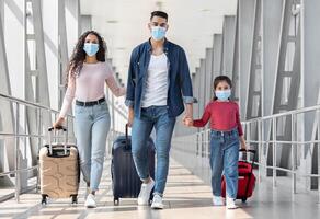 Travels During Pandemic. Arab Family Of Three Wearing Protective Medical Face Masks Walking With Suitcases At Airport Terminal, Arab Parents With Little Daughter Holding Hands While Going To Gate photo