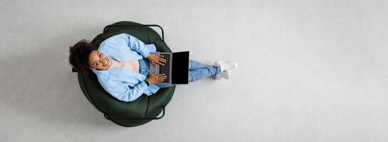 People And Modern Technology. Above top view of young smiling black woman holding pc on lap sitting on bean bag in living room, looking up at camera. Lady browsing internet, free copy space panorama photo