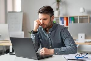 Serious middle aged businessman using laptop computer and reading reports, sitting at workplace in office. Business and entrepreneurship problems concept photo