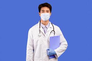 Portrait of serious asian male doctor wearing medical surgical mask, white uniform coat, stethoscope and protective gloves, holding folder, posing and looking at camera isolated over blue background photo