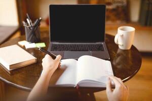 Note Taking Concept. Point of view of young woman sitting at table and writing, selective focus on notebook with pen. Lady using computer with blank empty screen, watching online course or webinar photo