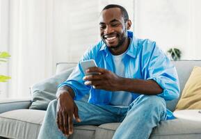 Cool gadget and app concept. Portrait of happy black man using mobile phone, watching while sitting on sofa in living room, free space. Guy browsing internet on cellphone photo