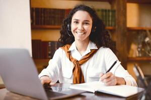 Remote Education During Quarantine. Portrait of cheerful woman sitting at table with pen, writing in notebook, using laptop and taking notes. Positive female student learning, looking at camera photo