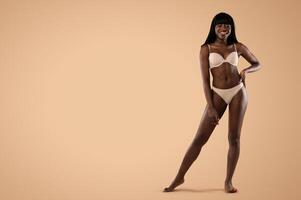 Happy attractive slender millennial african american lady posing in nude underwear over beige studio background, showing her beautiful curves, panorama with copy space, full length shot photo