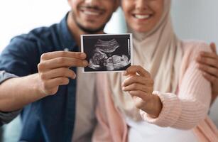 Closeup shot of happy pregnant muslim couple demonstrating baby sonography photo while relaxing together at home, showing first photo of their child, enjoying future parenthood, cropped image