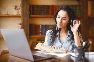Knowledge Concept. Portrait of calm young lady wearing and touching wireless headphones, reading paper book, sitting at desk with laptop computer, studying online at home, doing homework, taking break photo