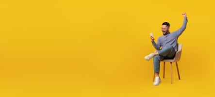 Asian man sitting on a chair beams with joy as he raises his fist in celebration while looking at his smartphone against a vibrant yellow backdrop. photo