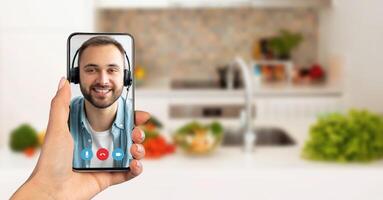 A person holds a smartphone showing a smiling individual on a call. The background features a cozy kitchen with fresh vegetables, highlighting a casual conversation atmosphere. photo