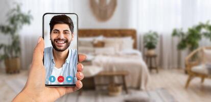 A person holds a smartphone showing a smiling face on a call. The background reveals a comfortable room decorated with plants and soft furnishings, creating a warm atmosphere for communication. photo