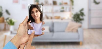 A person connects with someone through a smartphone during a call. The setting is a cozy home with a light-filled living room. The interaction appears warm and engaging. photo