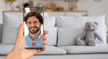 A person participates in a call, holding a smartphone with a smiling face on the screen. The setting is a cozy living room with a couch and a teddy bear, creating a warm atmosphere. photo
