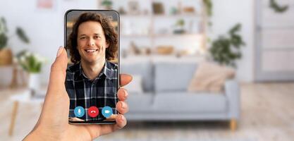 A hand holds a smartphone displaying a man smiling during a call. The setting features a cozy home interior with a modern couch and plants in the background, creating a relaxed atmosphere. photo