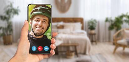 A person smiling while on a call appears on a smartphone screen. The background features a well-decorated home interior with light curtains and comfortable furniture. photo