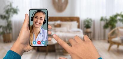 A person is using a smartphone to participate in a call, interacting with a smiling individual wearing a headset. The setting is a cozy home environment with soft furnishings. photo