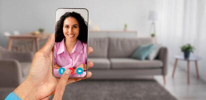 A person participates in a call using a smartphone, interacting with smiling woman from a cozy home environment. The setting includes a modern living room with comfortable furniture. photo