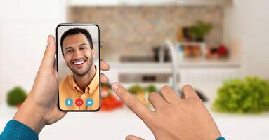 A person engages in a call using a smartphone in a bright kitchen, pointing at the screen with a smiling face of another individual visible. Fresh vegetables are placed in the background. photo