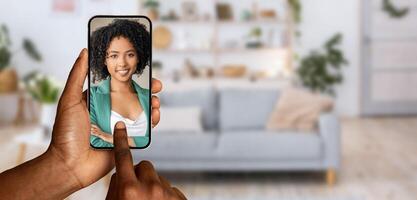 A person is holding a smartphone and participating in a call from their living room. The background features a cozy sofa and decorative elements, creating a relaxed atmosphere. photo
