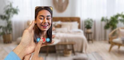 A person is holding a smartphone while having a call in a cozy room. The background features a comfortable bed and plants, creating a warm atmosphere for conversation. photo