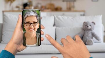 A person is using a smartphone for a call from their home. The lady on screen smiling and engaged in conversation while relaxing on a couch with a teddy bear nearby. photo