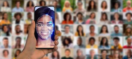 A person holds a smartphone displaying a face recognition scan in front of a collage of diverse faces. This showcases modern identification technology in action, highlighting its potential uses. photo