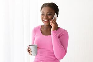 Excited Black Woman With Cellphone And Coffee Near Window Enjoying Phone Call, African American Lady Holding Cup With Hot Drink And Having Pleasant Mobile Conversation, Relaxing At Home, Copy Space photo
