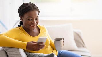 Weekend Leisure. Cheerful black female using smartphone and drinking tea at home, youg lady resting in living room with cellphone and coffee, browsing internet or checking social networks, copy space photo