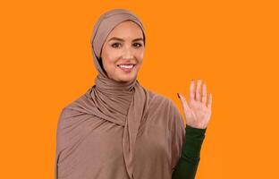 Hello Greeting. Muslim Lady In Hijab Waving Hand Showing Hi Gesture Smiling To Camera Standing On Yellow Studio Background. Cheerful Arabic Woman Gesturing Goodbye Concept photo
