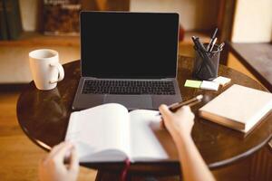 Online Class Concept. Point of view of female student sitting at desk and writing in notebook, taking notes, using laptop with black empty screen for mock up template, watching lecture or webinar photo