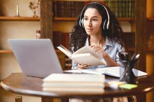 Virtual Lecture. Portrait of young lady in wireless headphones sitting at desk, holding and reading book, having online class with tutor and looking at laptop computer. Woman studying at home photo