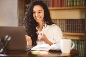 Online Call And Communication. Portrait of smiling curly woman sitting at table, using laptop and having virtual conference on computer, talking to webcam, explaining plan, selective focus photo