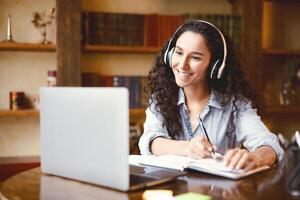 Home Office. Portrait of smiling young lady in wireless headphones sitting at desk, writing in notebook and using computer. Woman watching online tutorial, webinar or seminar, studying at home photo