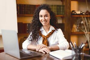 Portrait of positive casual curly young woman sitting at table and using laptop, bookshelves and board in the blurred background. Lady smiling and ready for online tutoring or call photo
