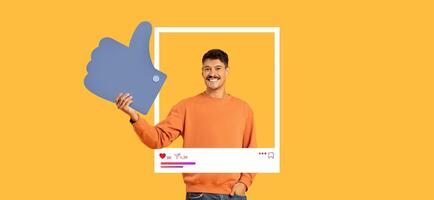 A man in an orange sweater expresses positivity by giving a thumbs up while holding a large like symbol. Bright yellow background enhances the cheerful vibe, perfect for social media engagement. photo