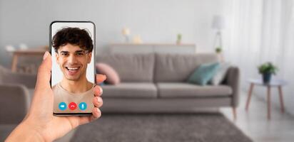 A person holding a smartphone during a call at home. The cozy living room features a sofa and soft lighting, creating a comfortable atmosphere for virtual interaction. photo