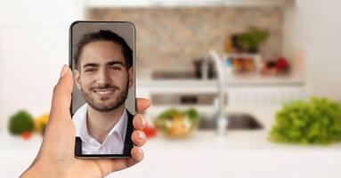 A person holds a smartphone displaying a smiling man during a call. The background features a modern kitchen with fresh vegetables and a bright atmosphere, indicating a casual home setting. photo