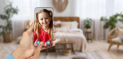 A person holds a smartphone displaying a smiling child during a call. The cozy home environment features a well-decorated living space with plants and soft furnishings. photo